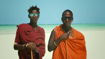 young masai tribe members in red dresses and sunglasses standing on sand beach of the indian ocean