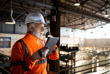 Industrial Engineer Worker Wearing Safety Uniform And Hardhat Standing On Metal Platform And Checking Production On Tablet Computer. Factory Interior.