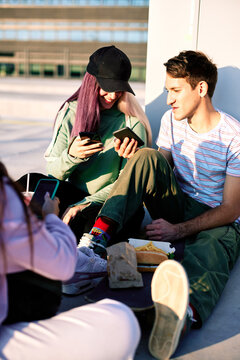 Happy teenage friends are sitting in urban exterior and using their phones for hanging on social media.