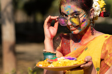 Wall Mural - Holi festival of color, young beautiful girl or woman or lady in indian attire saree holding a clay bowl full of dry organic color colour gulal abeer Holi powder and flower