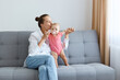 © sementsova321 - Horizontal shot of lovely woman with bun hairstyle wearing white shirt and jeans, young mother kissing her infant daughter while sitting on sofa and playing together.