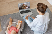 Back View Portrait Of Woman With Bun Hairstyle Wearing White Shirt And Jeans Sitting On Floor With Her Baby In Rocking Chair, Having Video Call On Laptop With Soldier Husband And Elder Daughter.