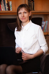 a female manager in a white shirt sits in a chair against the background of books with a laptop on her lap