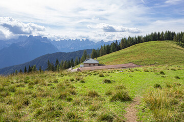 Wall Mural - Mountain summer landscape. Grassy meadow with a building and barn with the background of the forest and mountains under the sky with clouds. Cadore, Italy.