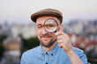 Cute male searching for sales or discounts at sunset using loupe with urban view. Attractive bearded man looking with magnifying glass at camera wearing jeans shirt and peaked cap. High quality image