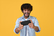 © insta_photos - Excited happy young indian man gamer player using smartphone playing game in mobile videogame app or watching digital video stream holding cell phone isolated on yellow background.