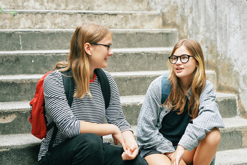 Outdoor portrait of two teenage girl sitting on stairs, wearing backbacks, talking to each other