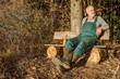 A farmerwith green dungarees and rubber boots relaxes after work on his bench at the edge of the forest in the mild evening sun.