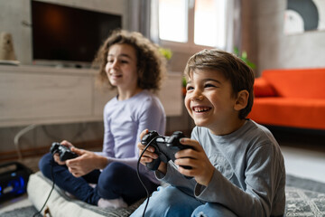 Poster - Two children small caucasian brother and sister happy children siblings boy and girl playing video game console using joystick or controller while sitting at home real people family leisure concept