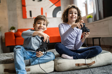 Wall Mural - Two children small caucasian brother and sister happy children siblings boy and girl playing video game console using joystick or controller while sitting at home real people family leisure concept