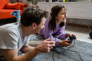 Canvas Print - Man and woman lying on the floor at home playing video game console having fun young couple man and woman spending time together holding joystick controller smiling on the floor at home real people