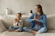 © fizkes - Happy mom and peaceful little daughter kid meditating at home together, sitting on couch with Namaste hands. Daycare teacher, mentor teaching preschool child to do yoga. smiling with closed eyes