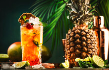 Alcoholic Cocktail With Vodka, Pineapple Juice, Mango, Red Syrup And Ice. Long Drink Or Summer Cold Mocktail. Tropical Dark Background With Palm Leaves And Exotic Fruits