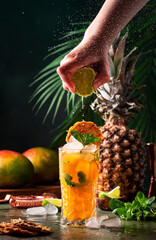 Wall Mural - Summer cocktail with vodka, pineapple juice, mango, ice. Long drink or cold mocktail. Bartender hand squeezes lime juice, frozen motion and flying drops. Tropical background with palm leaves