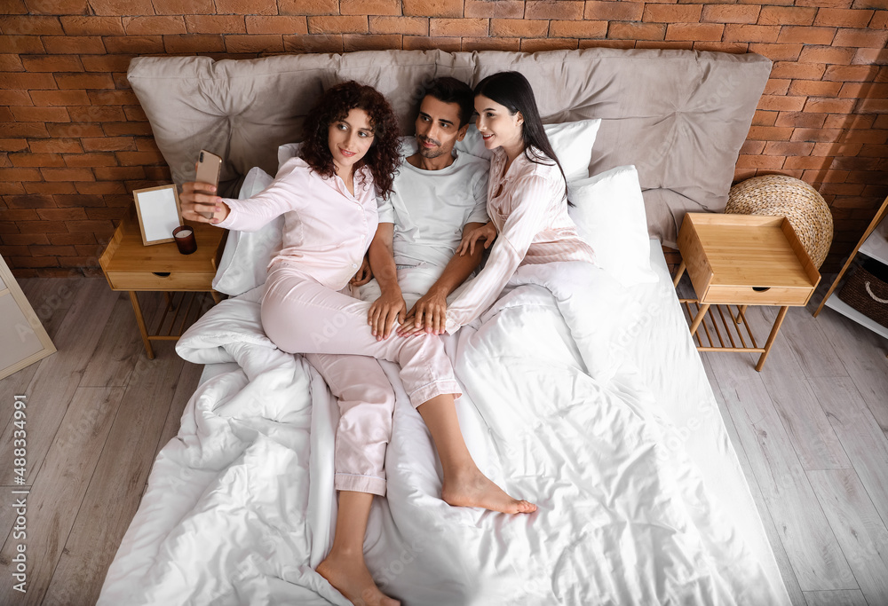 Man with two beautiful women taking selfie in bed. Polyamory concept