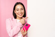 Image of smiling asian corporate woman in suit, holding smartphone, pointing at board, showing chart or information logo on empty white wall, standing over pink background