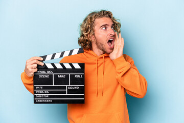 Wall Mural - Young caucasian man holding a clapperboard isolated on blue background shouting and holding palm near opened mouth.