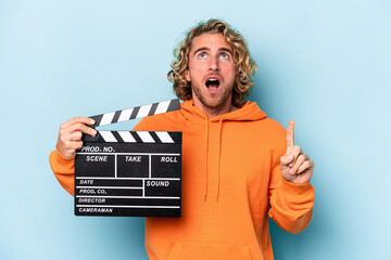 Wall Mural - Young caucasian man holding a clapperboard isolated on blue background pointing upside with opened mouth.