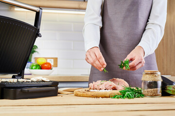 A female chef in a gray apron cooks or decorates raw meat with rosemary in the kitchen. Concept female cooks food at home.