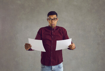 African American man with a puzzled and confused expression stands with two white sheets of paper. Young man in shirt holding blank layouts in left and right hand standing on gray background. Banner.