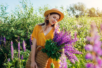 Woman put bouquet of lupin flowers in straw bag. Girl walking in summer meadow wearing yellow clothes.