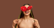 Portrait Of A Beautiful Woman On A Brown Background Holding Two Red Flowers Hiding Their Eyes And Laughing. Beauty Concept.