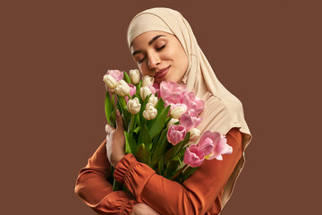 Wall Mural - Portrait of a beautiful muslim woman wearing a beige hijab and holding a big bouquet of tulips on a brown background. The concept of female beauty, spring and femininity.