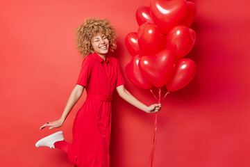 Wall Mural - Happy energetic curly haired woman has fun dances and raises leg expresses positive emotions holds bunch of inflated heart shaped helium balloons comes on party isolated over vivid red background.