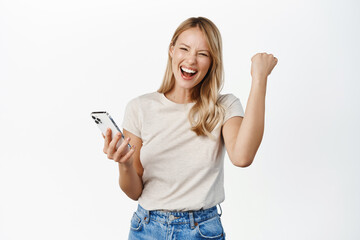 Wall Mural - Enthusiastic young woman winning on mobile phone, rejoicing, using smartphone app, celebrating, triumphing on cellphone, white background