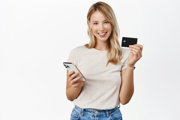 Wall Mural - Beautiful smiling woman showing credit card while paying with mobile phone app, using smartphone, shopping online, purchase smth,white background