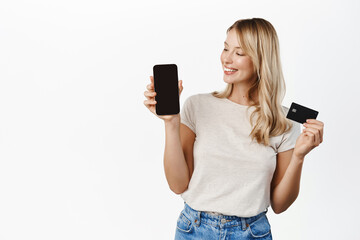 Wall Mural - Beautiful girl smiling, showing smartphone screen and credit card, paying, shopping application app interface, standing over white background