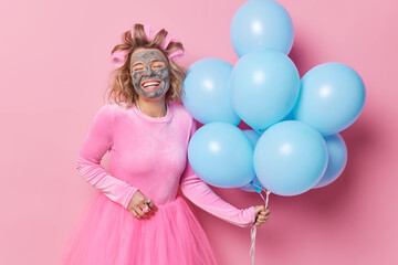 Wall Mural - Happy woman smiles broadly undergoes beauty procedures applies clay mask and hair rollers wants to have perfect outlook wears dress holds bunch of blue balloons prepares for party. Celebration