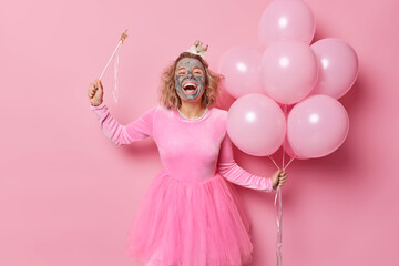 Wall Mural - Positive woman prepares for birthday celebration applies beauty mask on face to look beautiful holds magic wand and bunch of helium balloons wears dress poses against pink background. Holidays