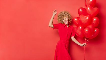 Wall Mural - Energetic woman with blonde curly hair wears dress dances carefree with bunch of heart shaped balloons has fun celebrates holiday isolated over red background with copy space for promo. Valentines Day