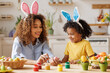 © JenkoAtaman - Happy african american family: mother teaching happy little kid soon to decorate Easter eggs while sitting in kitchen