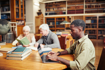 Side view portrait of young black man studying at table in classic college library with people in background