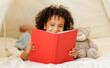 © JenkoAtaman - Cute little african american kid curly boy holding book lying in play tent with teddy bear toy