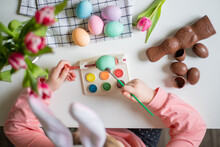 Top View Of A Little Girl With Bunny Ears Holding Paintbrush And Decorating Easter Eggs With Watercolors. Tulips, Easter Chocolate And Eggs On A White Table