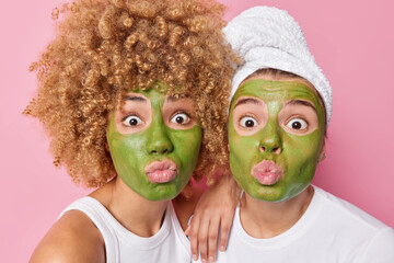 Wall Mural - Photo of surprised two women keep lips folded apply green nourishing masks take care of complexion and skin stand next to each other isolated over pink background. Beauty and wellness concept