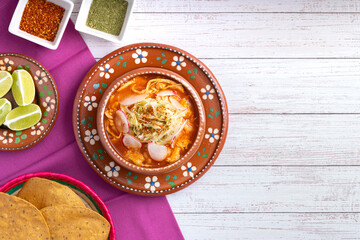 Traditional mexican food. Red pozole soup with chicken accompanied with avocado, lettuce, onion, radish, lemon, chili and crispy corn tortillas also known as tostadas on a white wooden background.