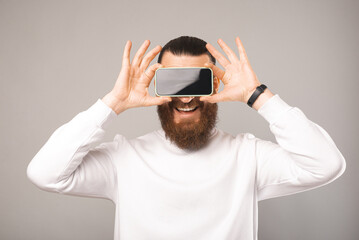 Young bearded man is covering eyes with his smartphone. Studio shot.