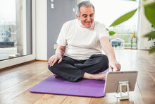 Smiling Senior Man Sitting Cross-legged On Exercise Mat Using Tablet PC At Home