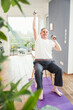 © Westend61 - Smiling senior man with hand raised holding dumbbell on chair at home