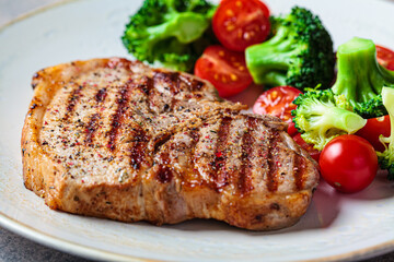 Canvas Print - Grilled pork steak with broccoli and tomatoes on plate, dark background.