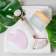 Facial Kit For Home Skincare And Spa. Rose Quartz Face Roller, Gua Sha Massagers, And Jar Of Face Cream Onwhite Towel Background With Green Monstara Leaves. Natural Treatment Concept. Top View.