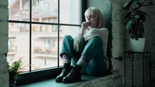 Beautiful Young And Blonde Caucasian Female Feeling Depressed And Biting Her Nails While Looking Worried. Sad Girl Sitting By The Window Crying By Herself.