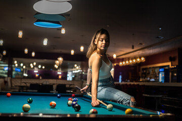 Wall Mural - Glamorous brunette woman holding a cue and sitting on the billiard table