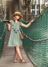 Portrait Of A Beautiful Caucasian Child Wearing A Straw Hat And A Green Dress In The Forest Standing In A Bridge Reading A Book

