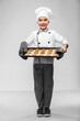 cooking, culinary and profession concept - happy smiling little boy in chef's toque and jacket holding baking tray with oatmeal cookies over grey background