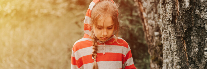 portrait face of upset or focused thoughtful scowl eight year old kid girl in hood of striped hoodie in the forest nature. prepubertal children mind psychology and mental health. banner. flare